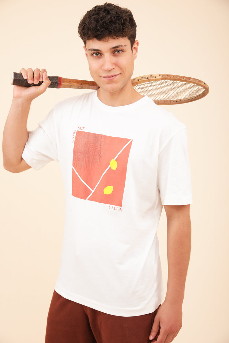 Homme en tshirt LYOUM sport terrain de tennis ocre et raquette à la main.
