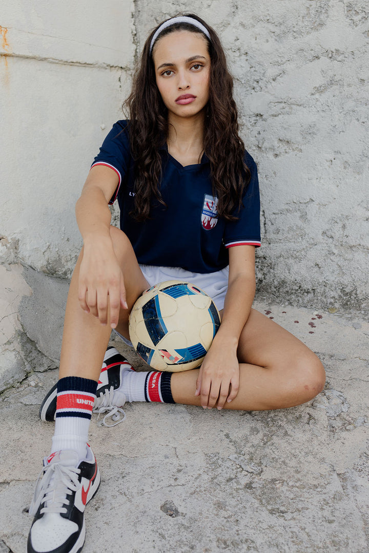 Femme en bandeau et maillot LYOUM Harissa United bleu tenant un ballon de football.