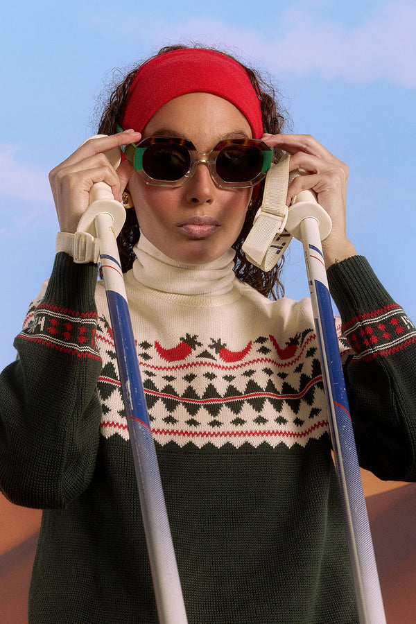 Femme en pull de noël LYOUM piment, lunettes de soleil et skis.