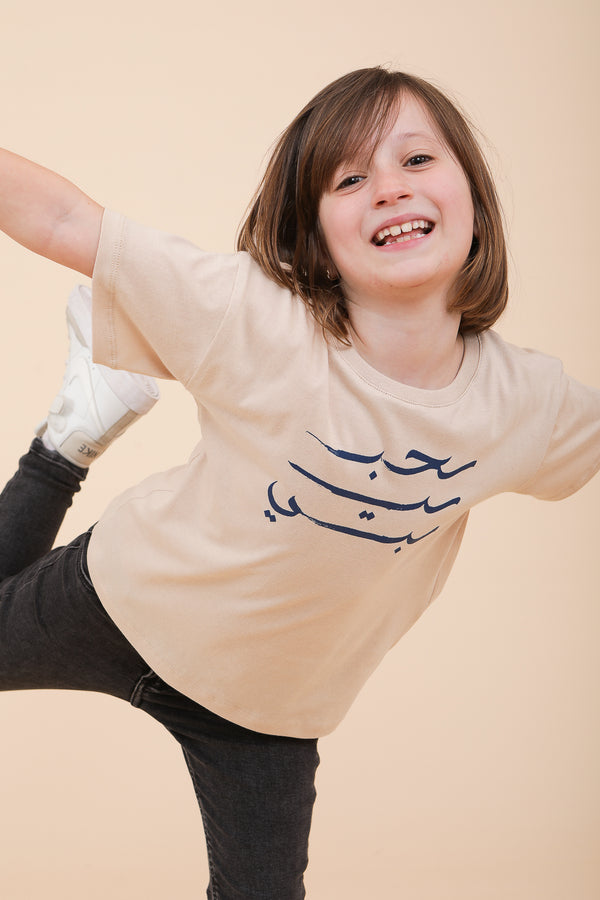 Petite fille souriante portant un t-shirt LYOUM en coton beige avec sérigraphie 'habibi' en arabe.