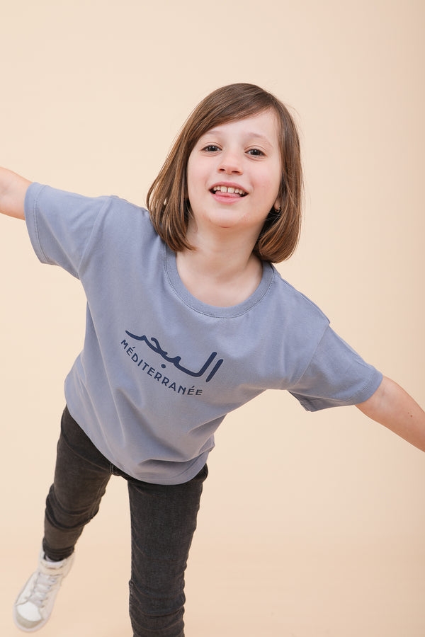 Petite fille portant un t-shirt LYOUM en coton bleu ciel avec sérigraphie 'La Mer Méditerranée' en arabe et français.